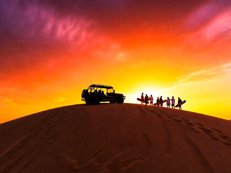 Grupo de turistas disfrutando el atardecer mágico (sunset) en la cima de las dunas de Paracas con tablas de sandboarding y tubular.