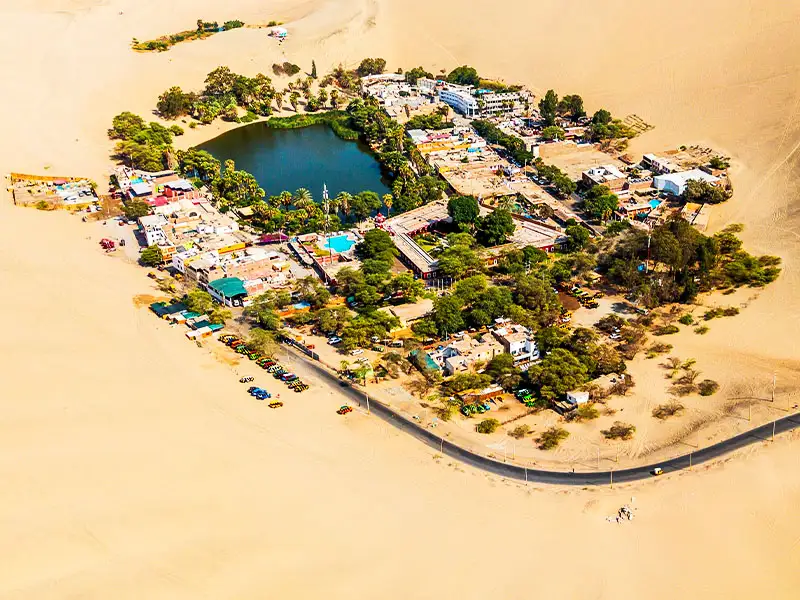Vista aérea panorámica del Oasis de Huacachina rodeado por el inmenso desierto de Ica, un destino popular para tours de aventura y tubulares areneros.