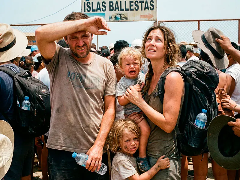 Fotografía documental de una familia exhausta y sudada esperando en una larga cola caótica bajo el sol en el muelle de Paracas debido a tours económicos.