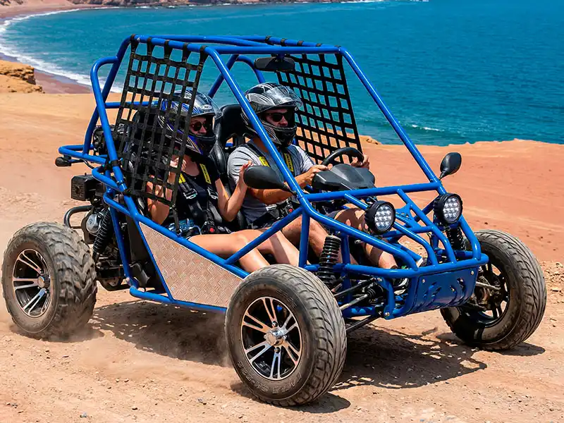 Pareja conduciendo un mini-buggy azul moderno y seguro por la ruta escénica de la Reserva Nacional de Paracas frente a la Playa Roja.