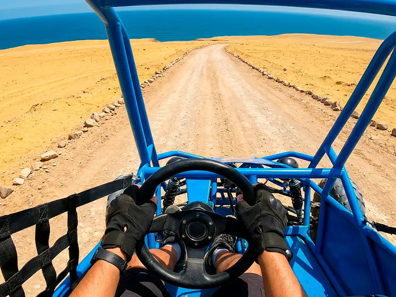 Vista en primera persona desde el interior de un mini-buggy azul, con las manos en el volante y el cinturón de seguridad puesto, conduciendo por un camino marcado y seguro en la Reserva Nacional de Paracas.