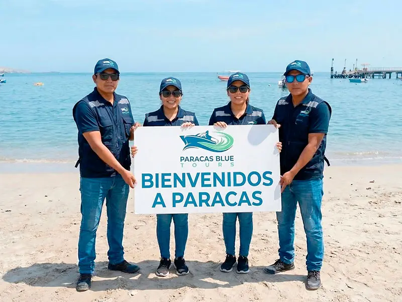 Equipo oficial de Paracas Blue Tours dando la bienvenida a los turistas en la playa de Paracas con cartel de seguridad.